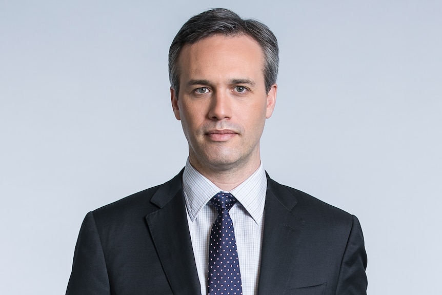 A middle aged man with a black suit, white shirt and a navy patterned tie.