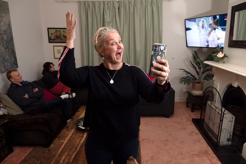 A woman stands excitedly in the centre of a room holding a  phone with people and a TV in the background