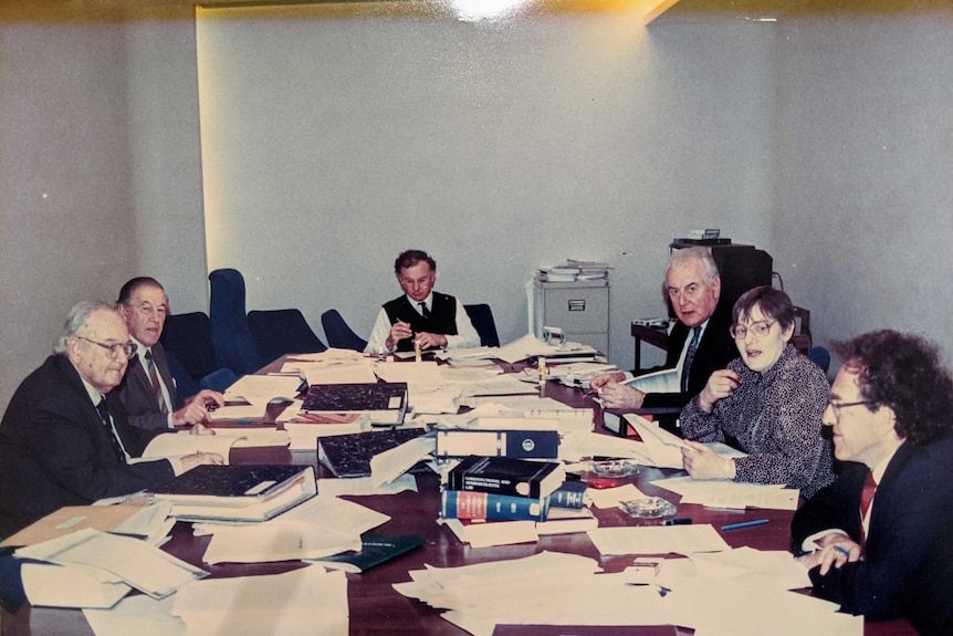 An old photo with six people sitting around a desk with books on it