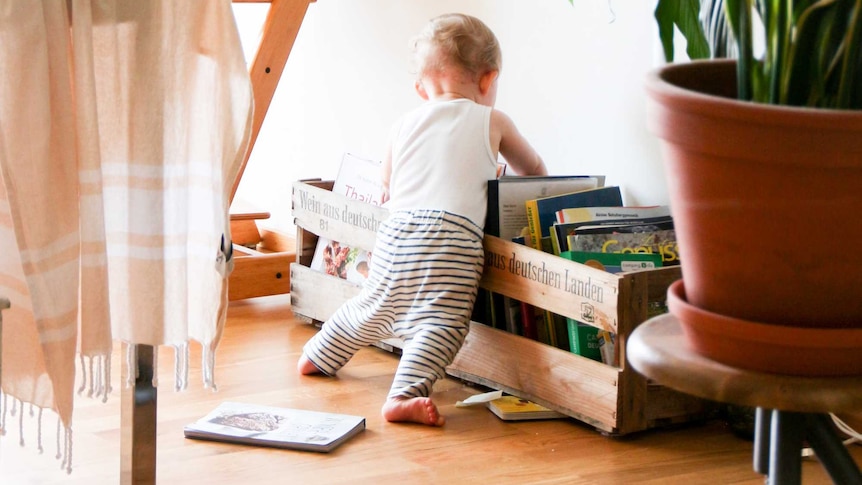 a very young child stands in a sunny room pulling itself up on a pile of books stacked on the floor