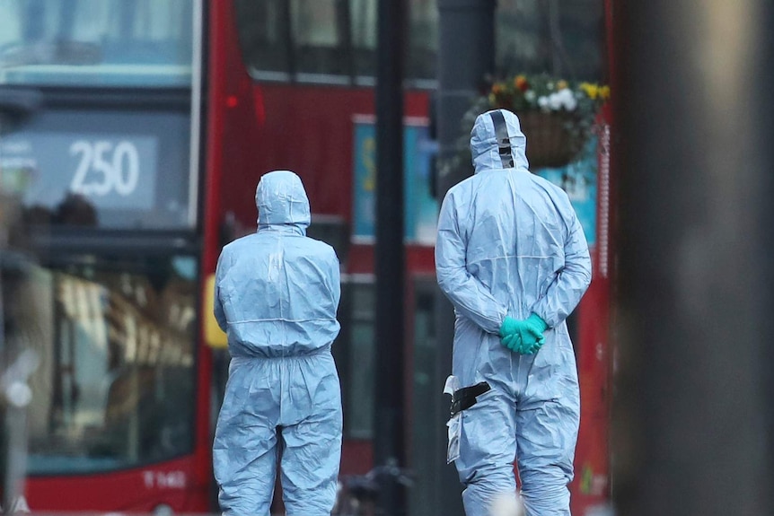 Two figures in forensic suits covering their bodies and gloves stand with their backs to the camera on a London street.