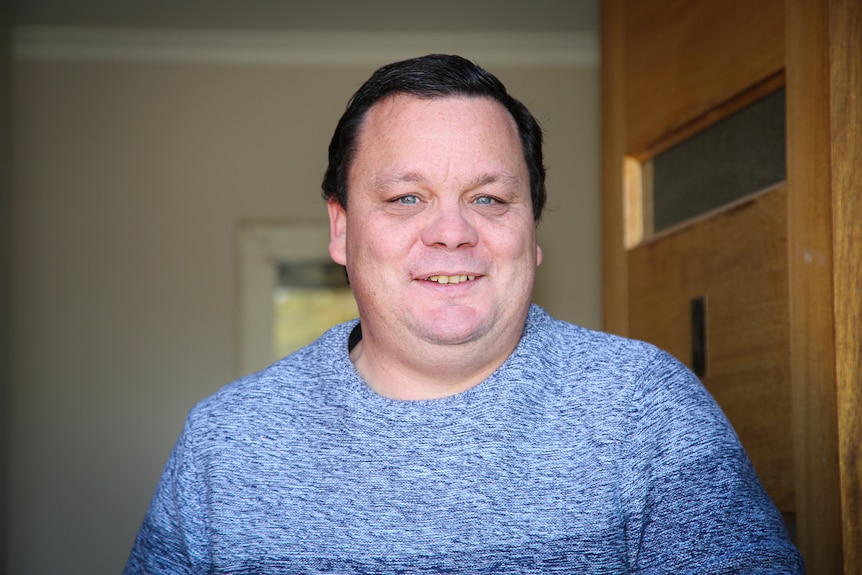 A man with short hair smiles while standing in his front doorway.