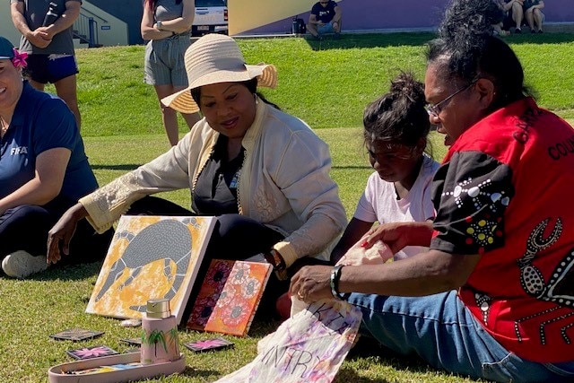 A hat-wearing senior FIFA executive sits on the grass and talks to two Indigenous women as they look at paintings..
