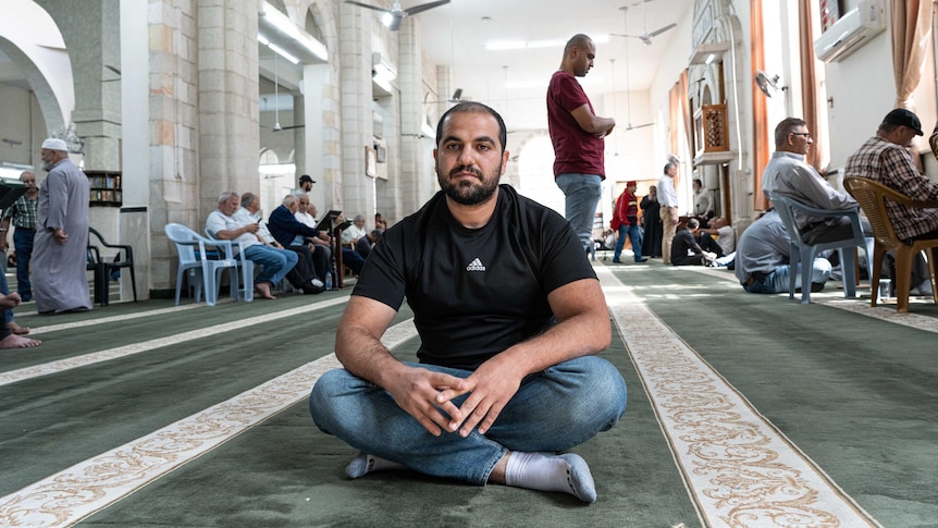 A man wearing jeans and a black T-shirt sits cross-legged on the floor of a mosque