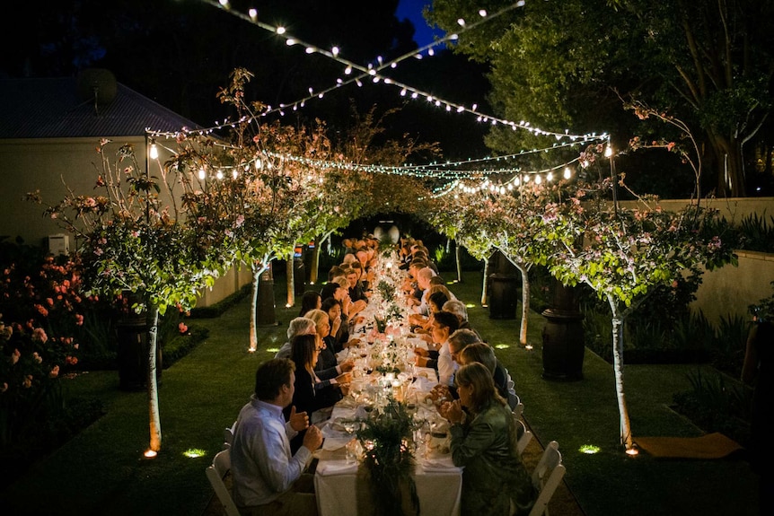 A long dinner table filled with guests at night in a garden under fairy lights.