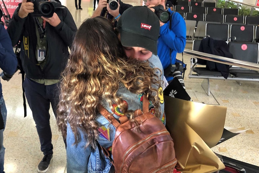 Two people embrace at the airport, one with long hair and the other in a cap