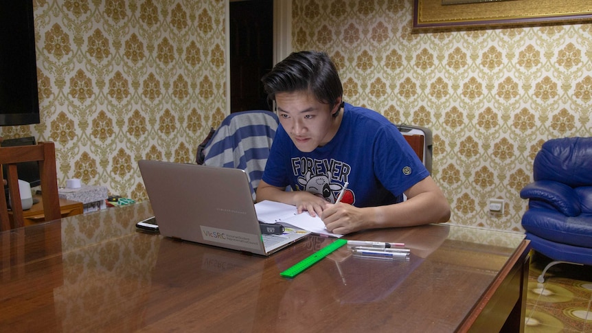 James Lam looks at his PC and takes notes in an exercise book while sitting at a dining room table