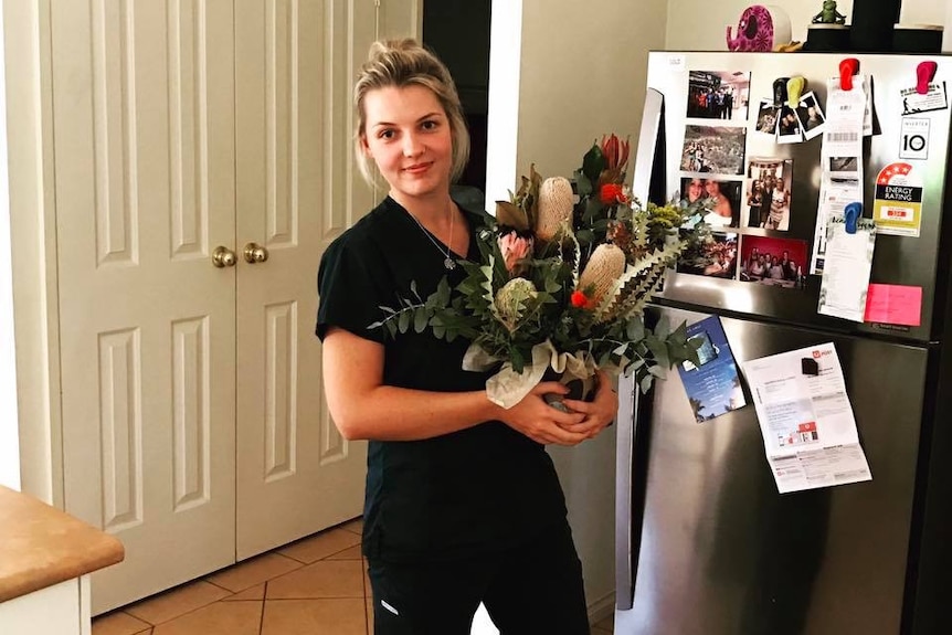 A woman in a nurses uniform holds a bunch of flowers in her home kitchen, after a tiring day at work.