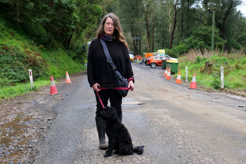 A woman wearing black stands on a country road with her black dog.