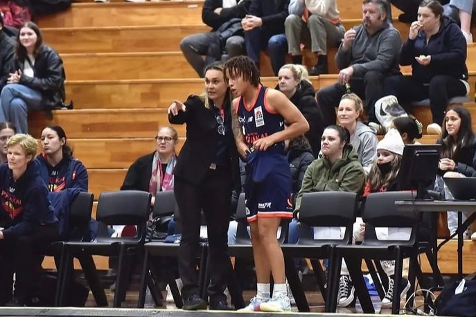 Tully Bevilaqua stands on the sideline of a basketball court with a player and points.