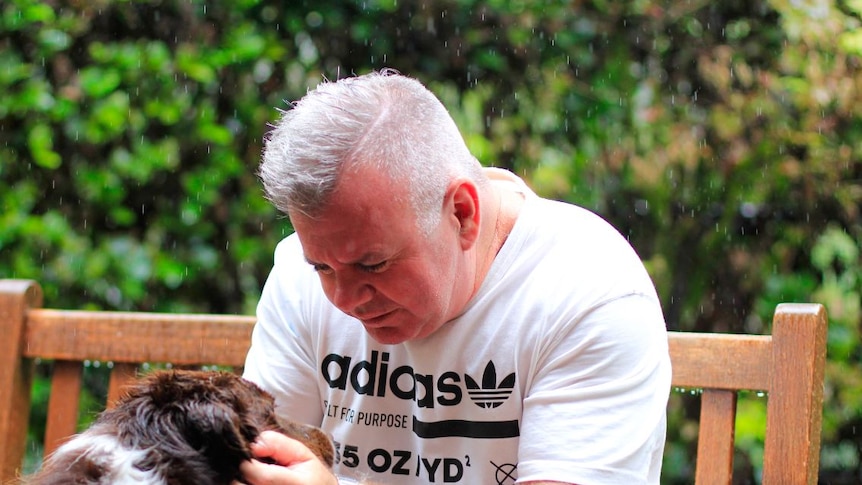 A grey haired man in a t-shirt sitting on a wooden bench patting a long haired dog