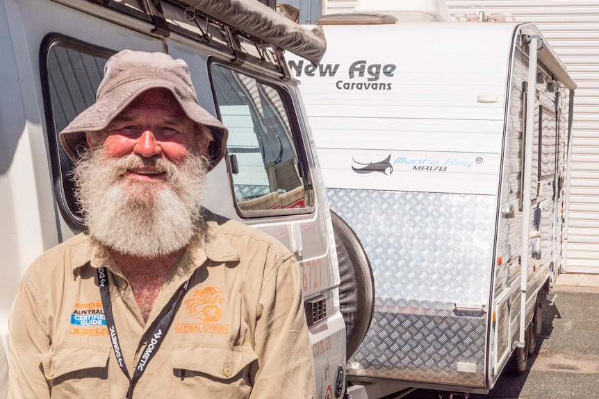 A man wearing a floppy hat stands in front of a four-wheel drive and caravan