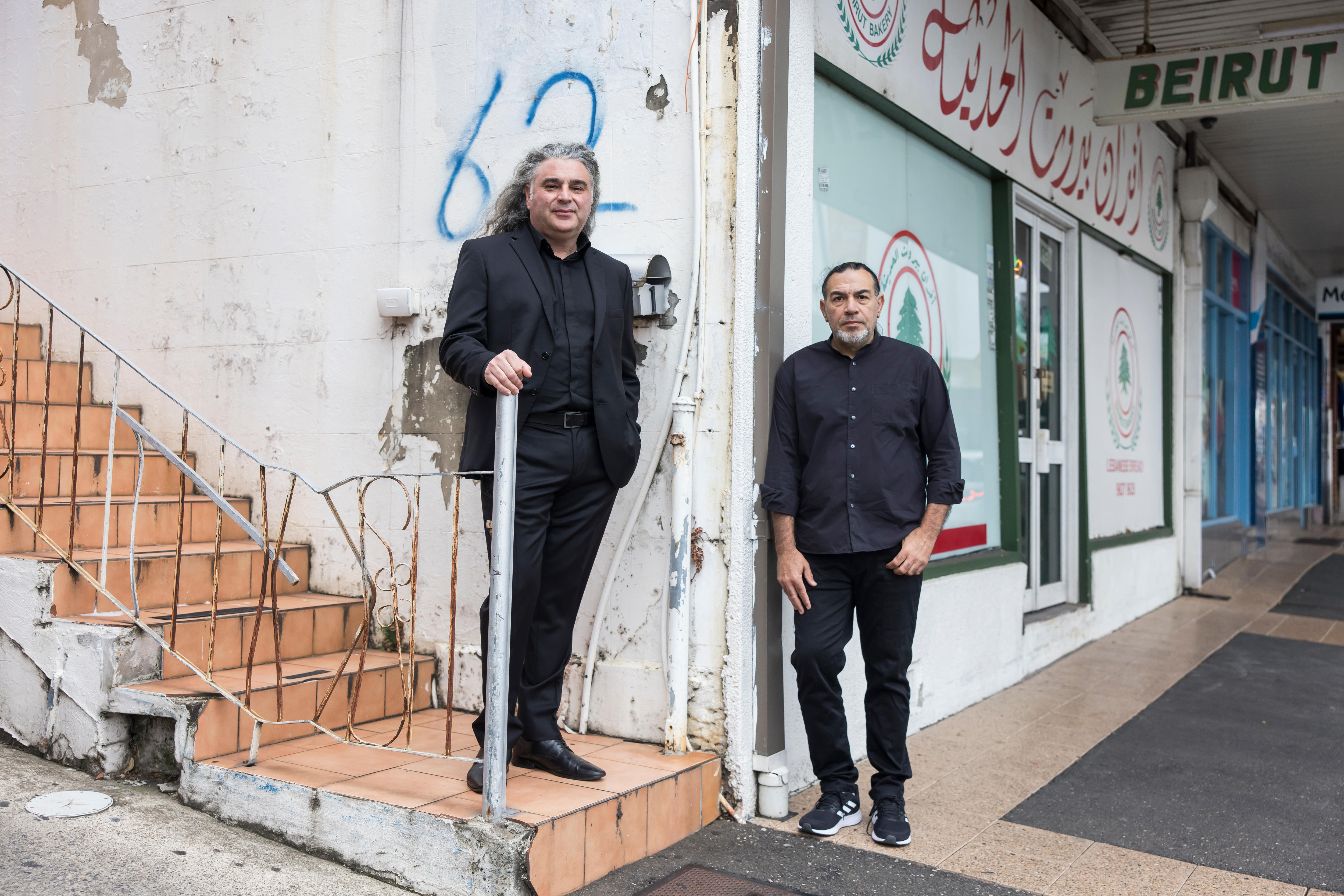 Two man stand on street wearing black suits. One smiles and one has serious expression.