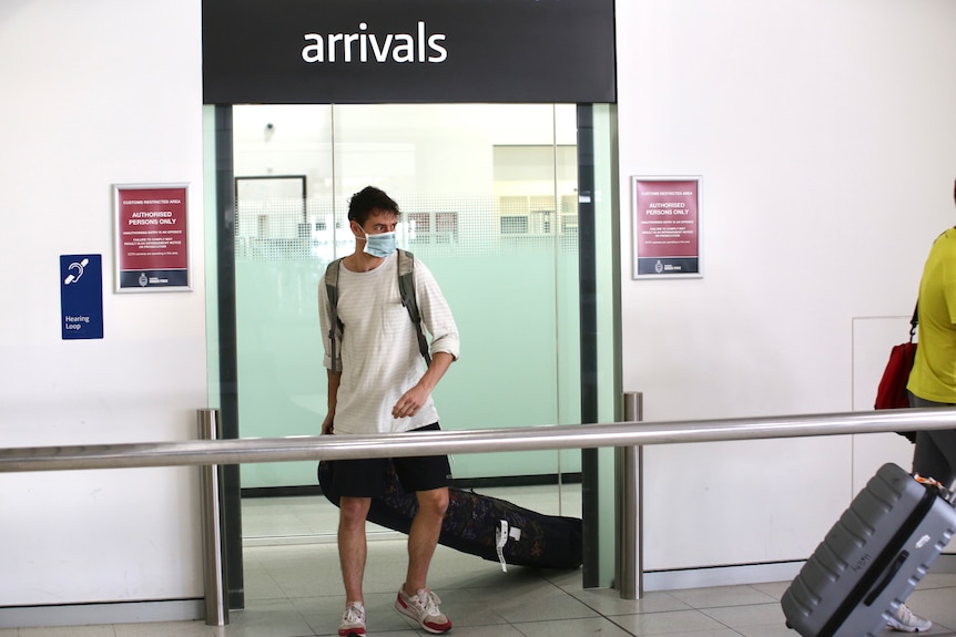 A young man wearing a blue face mask walks through the arrival gate at Perth Airport with luggage