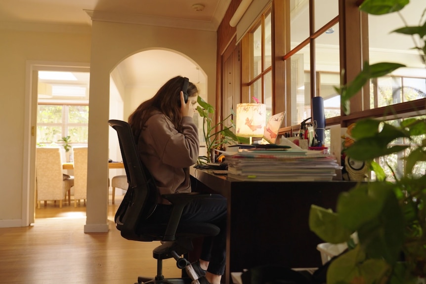 A young teenage girl wearing a brown jumper sits at a desk inside a house with books and a computer while putting headphones on.