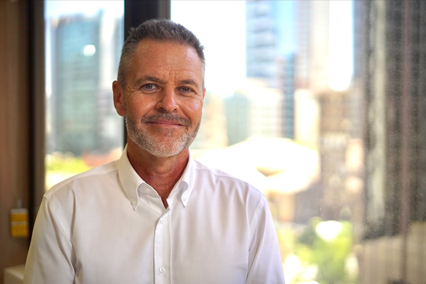 A smiling man wearing a white buttoned shirt stands in front of a window showing city buildings