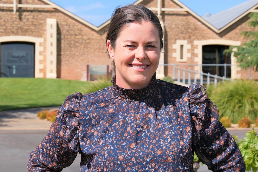 A middle aged woman smiles. It's a sunny day, she's standing in front of an old, well preserved brick building  