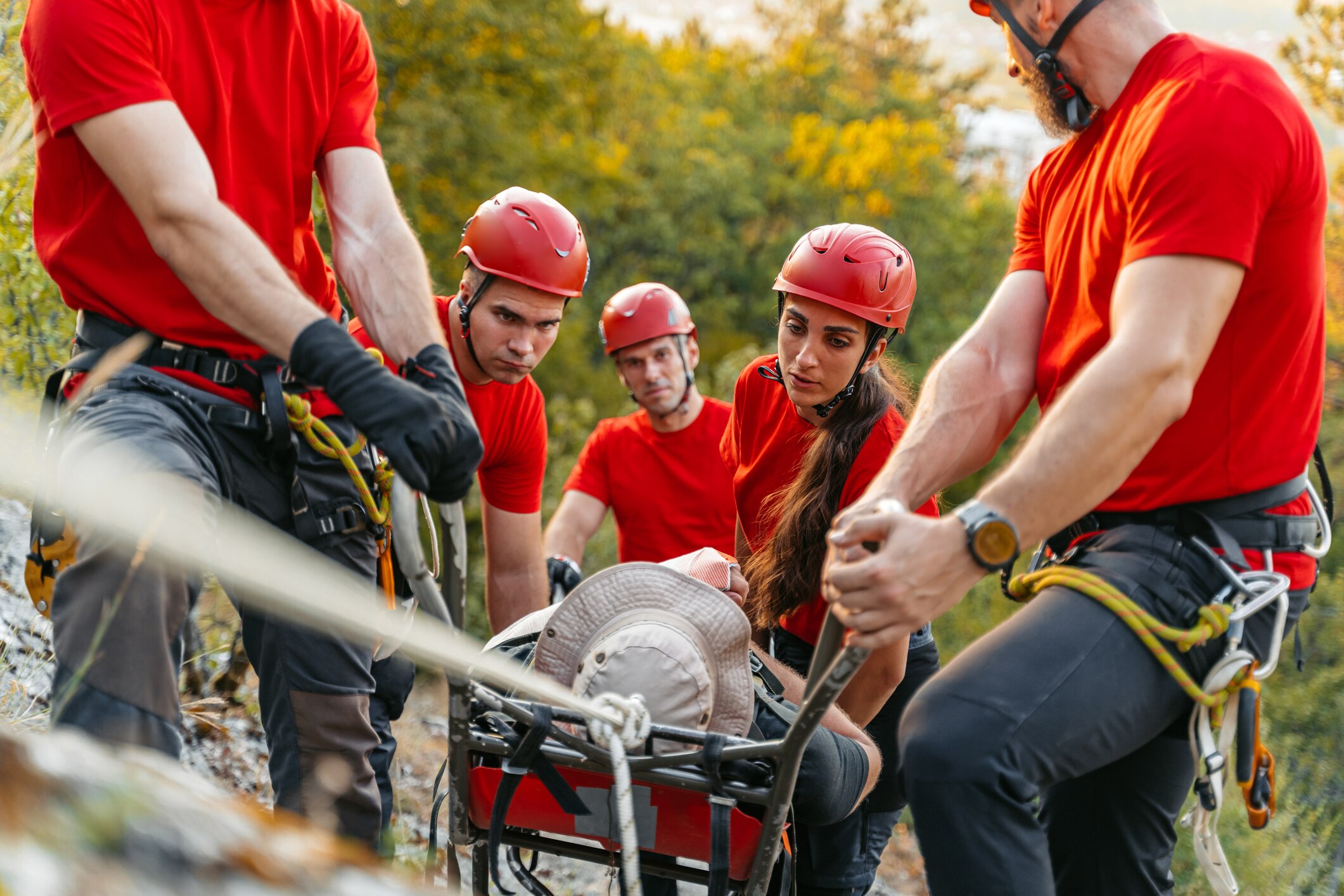 Rescuers wearing helmets and climbing ropes carry an injured male hiker on a stretcher up a steep rocky climb.