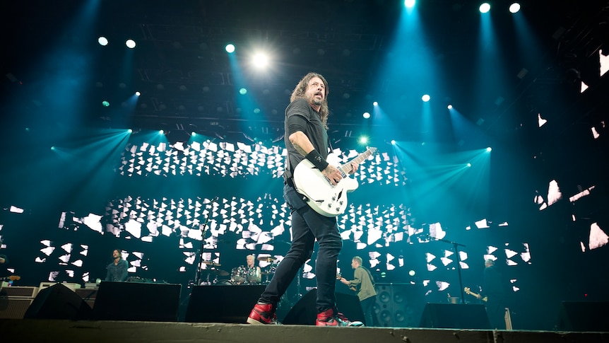 Dave Grohl stands up front of a rock show with his white guitar surveying the audience with lights and screens behind him