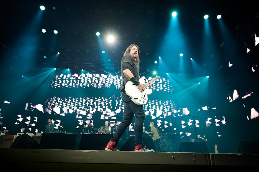 Dave Grohl stands up front of a rock show with his white guitar surveying the audience with lights and screens behind him