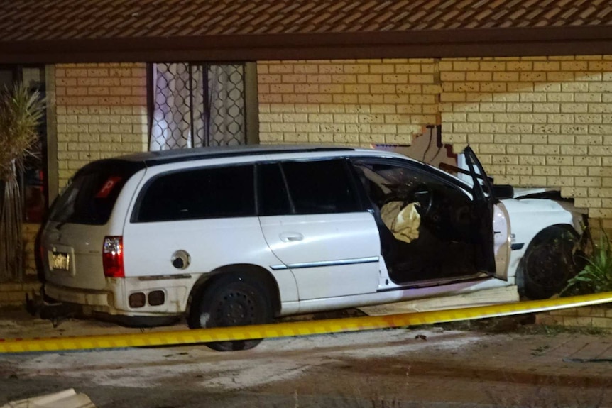 A white station wagon sticks out from the wall of a house after crashing into it.