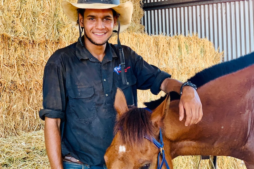 Image of an Aboriginal man standing with a horse in front of some bales of hay.