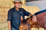 Image of an Aboriginal man standing with a horse in front of some bales of hay.