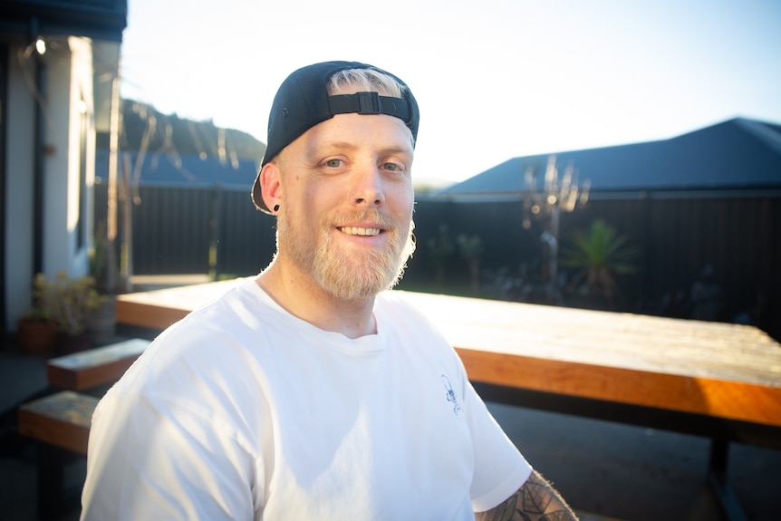 A man with a blonde beard wearing a white tshirt and a backward cap sitting in front of a window