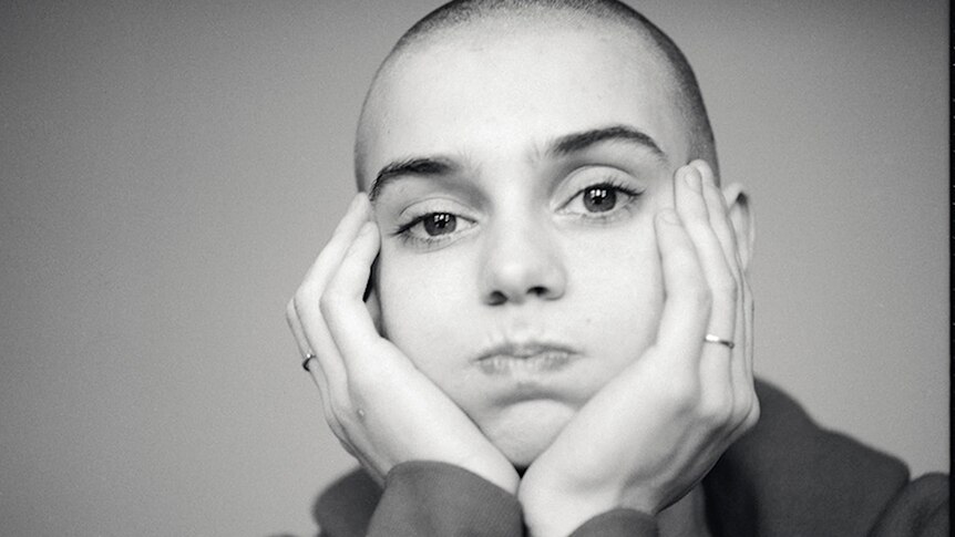 Black and white photo of a bald Sinead O'Connor puffing out her cheeks with her head in her hands as she looks at the camera