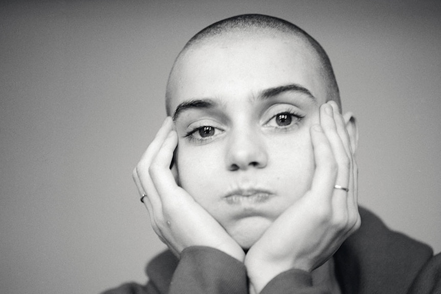 Black and white photo of a bald Sinead O'Connor puffing out her cheeks with her head in her hands as she looks at the camera