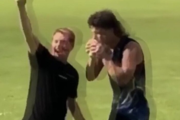 A man in a football uniform on grass drinks a drink with someone cheering next to him