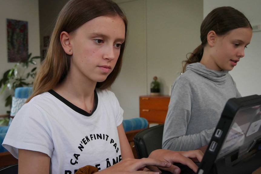 Tameka Brown in a white t-shirt and long hair working on her computer at the kitchen table, her younger sister in the background