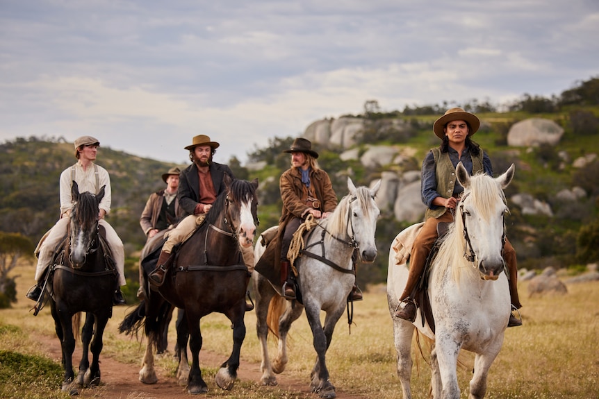 A group of men plus an Aboriginal woman all in 19th century clothing riding on horses in the bush