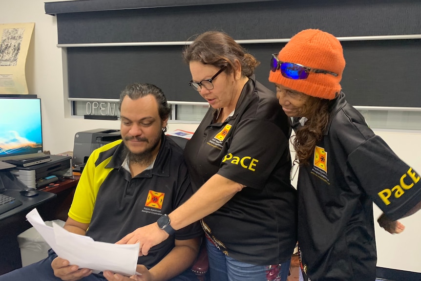 Josebessgo Mayers sits at a desk looking at a piece of paper handed to him by two other Aboriginal staff.