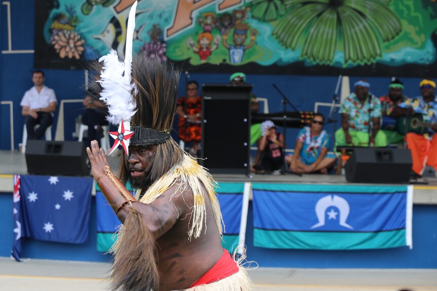 An Islander man wearing elaborate head gear, a grass skirt and feather decorations dancing