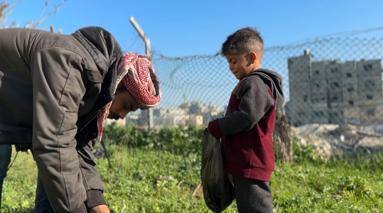 A man bends down to pull out weeds from the ground as a little boy watches on holding a black plastic bag.