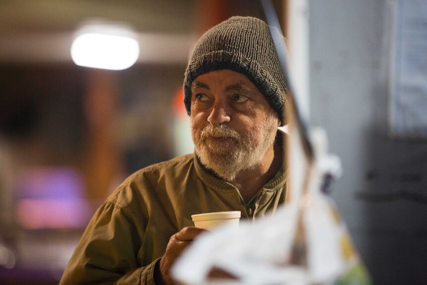 A man with a white beard and a beanie on holds a coffee cup.