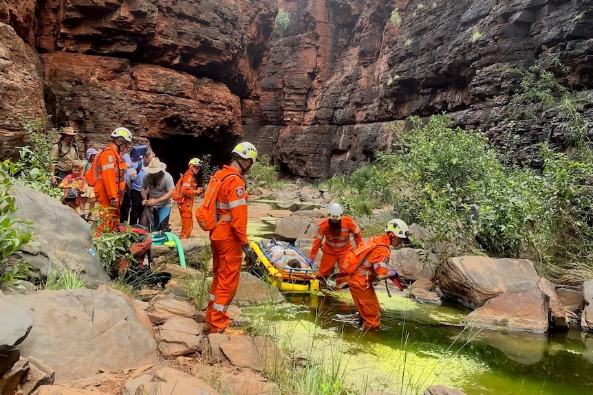 Emergency services crew carry a woman on a stretcher through a mossy lake in a gorge.
