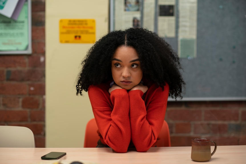 A Black woman in her late 20s with curly hair and red top leans on her hands looking concerned sitting at a meeting table