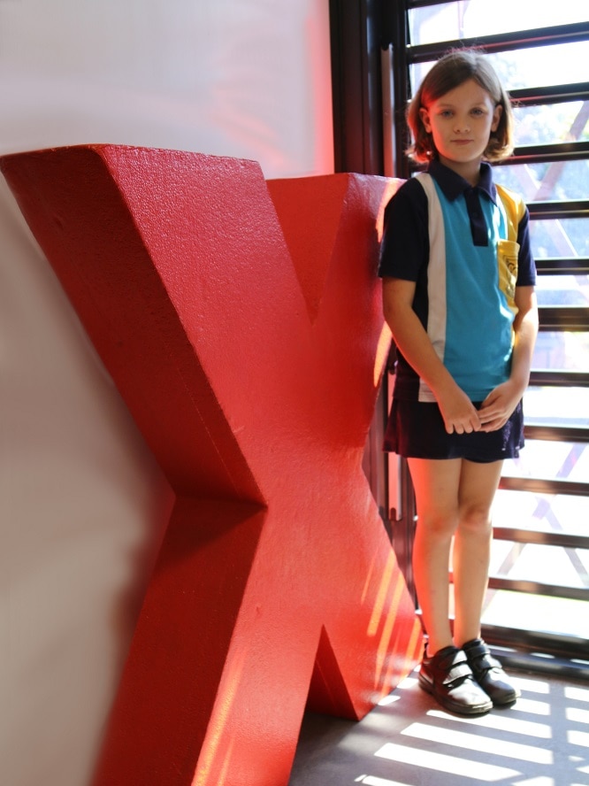 Nine-year-old Molly Steer in her school uniform standing next to a TedX sign and smiling.
