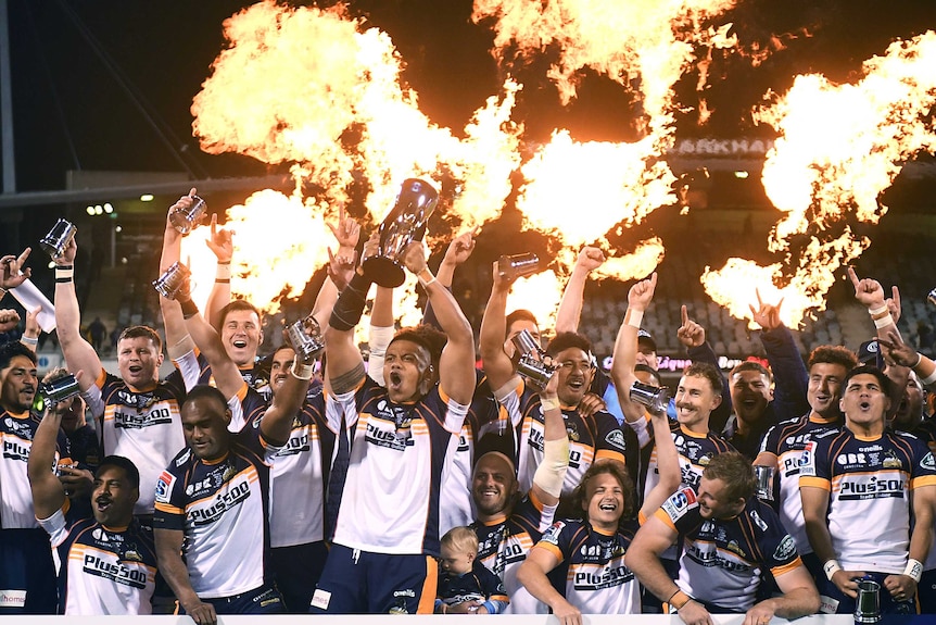 Rugby players wearing white shirts lift the Super Rugby AU trophy in the air, with flame throwers going off behind them