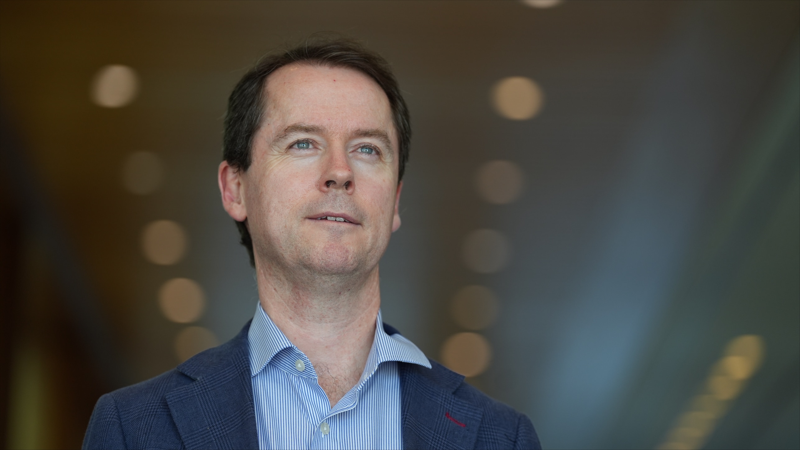 A man with brown hair wearing a blue button up shirt and navy jacket looks up and ahead.
