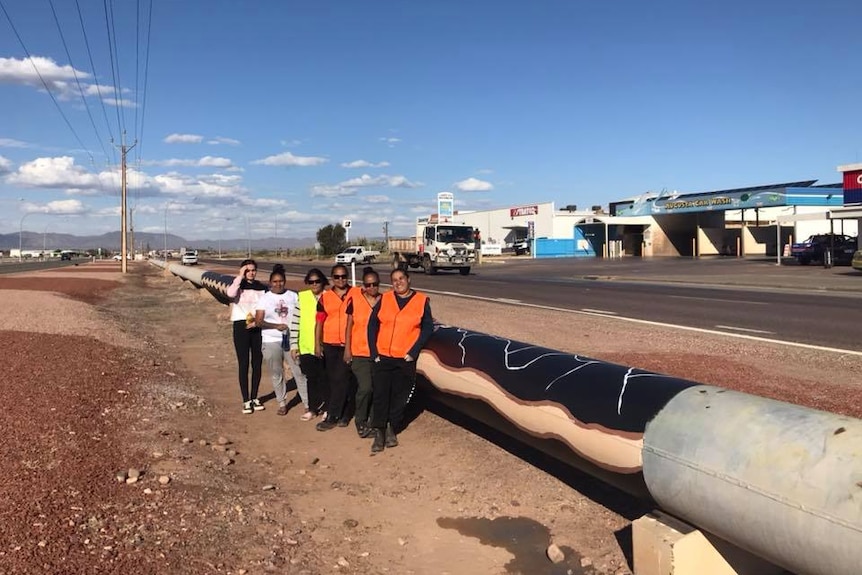 Three Aboriginal women wearing bright orange vests stand in front of a large pipeline which is painted black with white cracks.