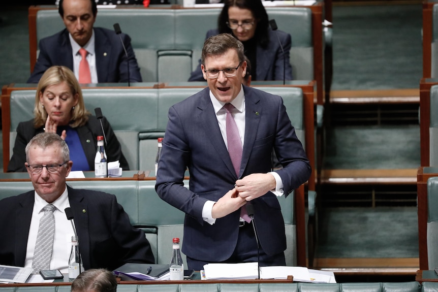 Tudge is standing, closing a jacket button, and talking with colleagues nearby.