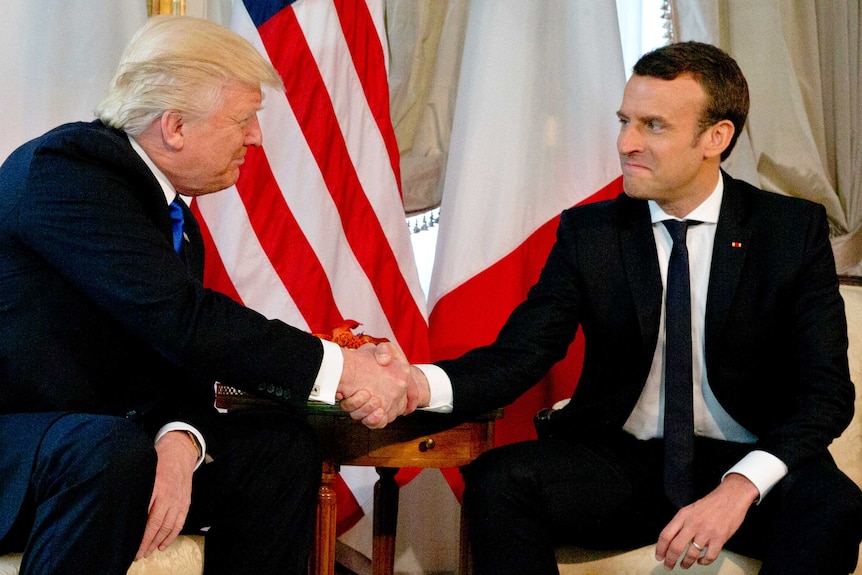 Donald Trump and Emmanuel Macron shakes hands and look at each other.