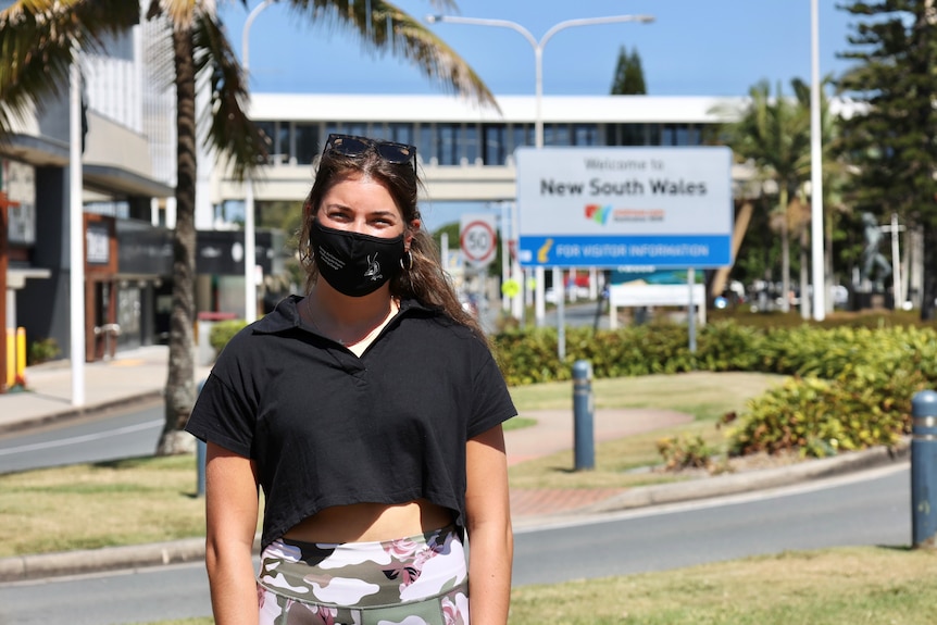 A masked young woman with dark hair and dark clothes stands in a regional town on a sunny day.