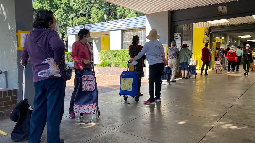 A line of people with their backs to the camera wait in line in a concrete corridor. 