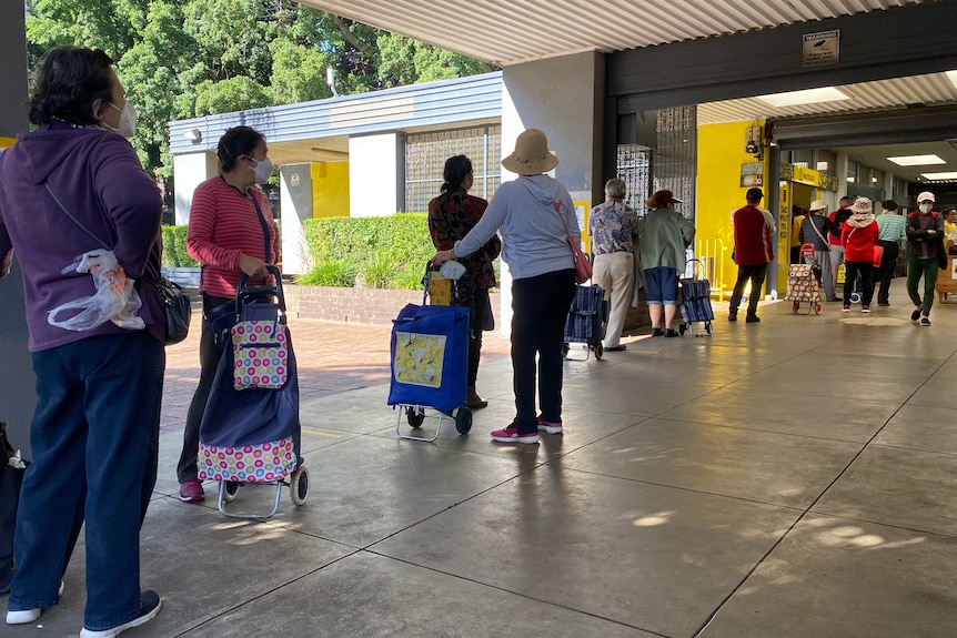 A line of people with their backs to the camera wait in line in a concrete corridor. 