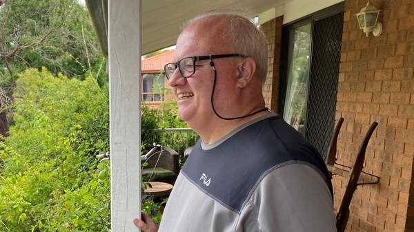 A man in a grey shirt stands on a verandah.