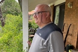 A man in a grey shirt stands on a verandah. 